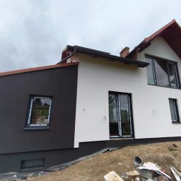 Tomasz Trojnar - Dwukolorowa elewacja domu z czarną i białą częścią. Widoczne okna z czarnymi ramami, drabina i fragment pracownika. Tło: pochmurne niebo.