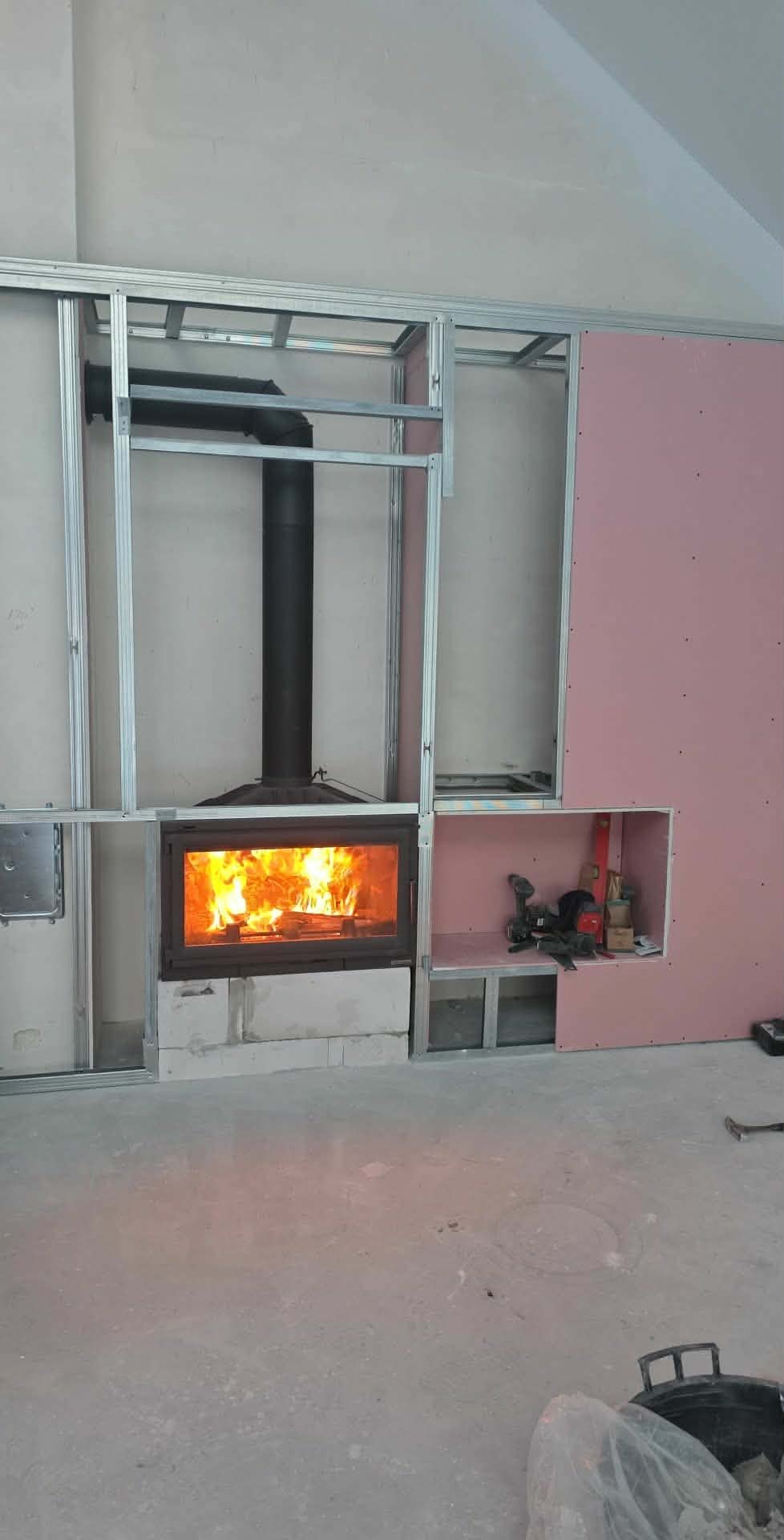 Wnętrze w trakcie wykończenia z widocznym kominkiem z paleniskiem, obudowanym płytami gipsowo-kartonowymi na metalowej konstrukcji. Widoczne narzędzia i elementy instalacji.