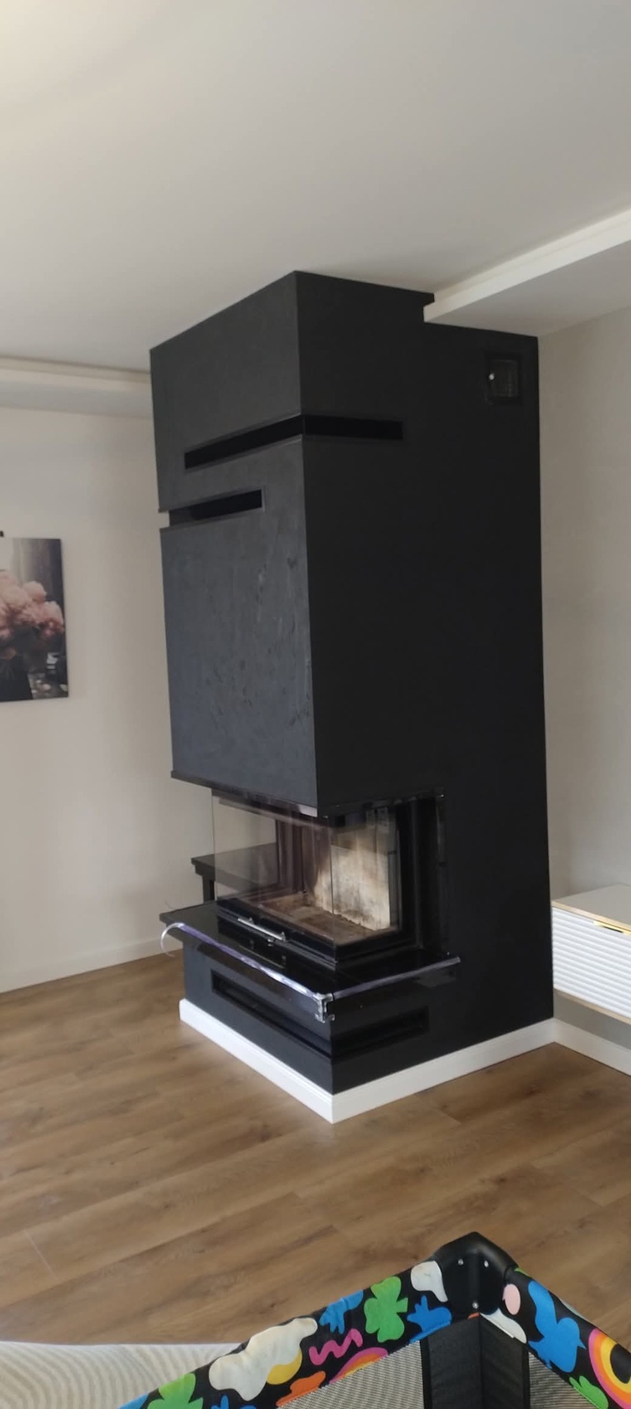 Nowoczesny, czarny kominek z przeszklonym paleniskiem, w jasnym wnętrzu z drewnianą podłogą. Minimalistyczny design, widoczne detale wykończenia.