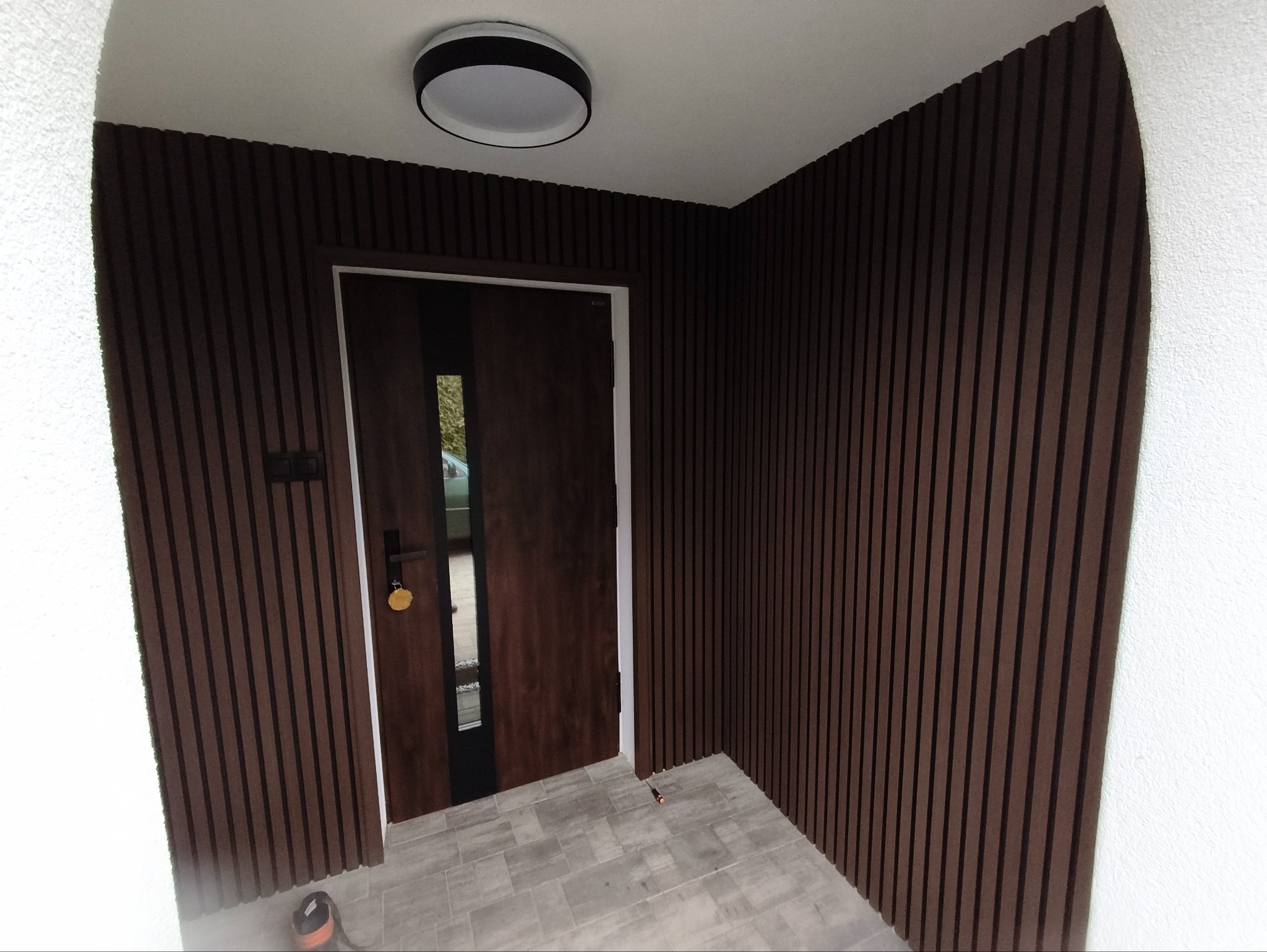 Nowoczesne wykończenie wnętrza: brązowe panele ścienne z pionowymi listwami, białe ściany, szara podłoga i drewniane drzwi z czarnym akcentem.
