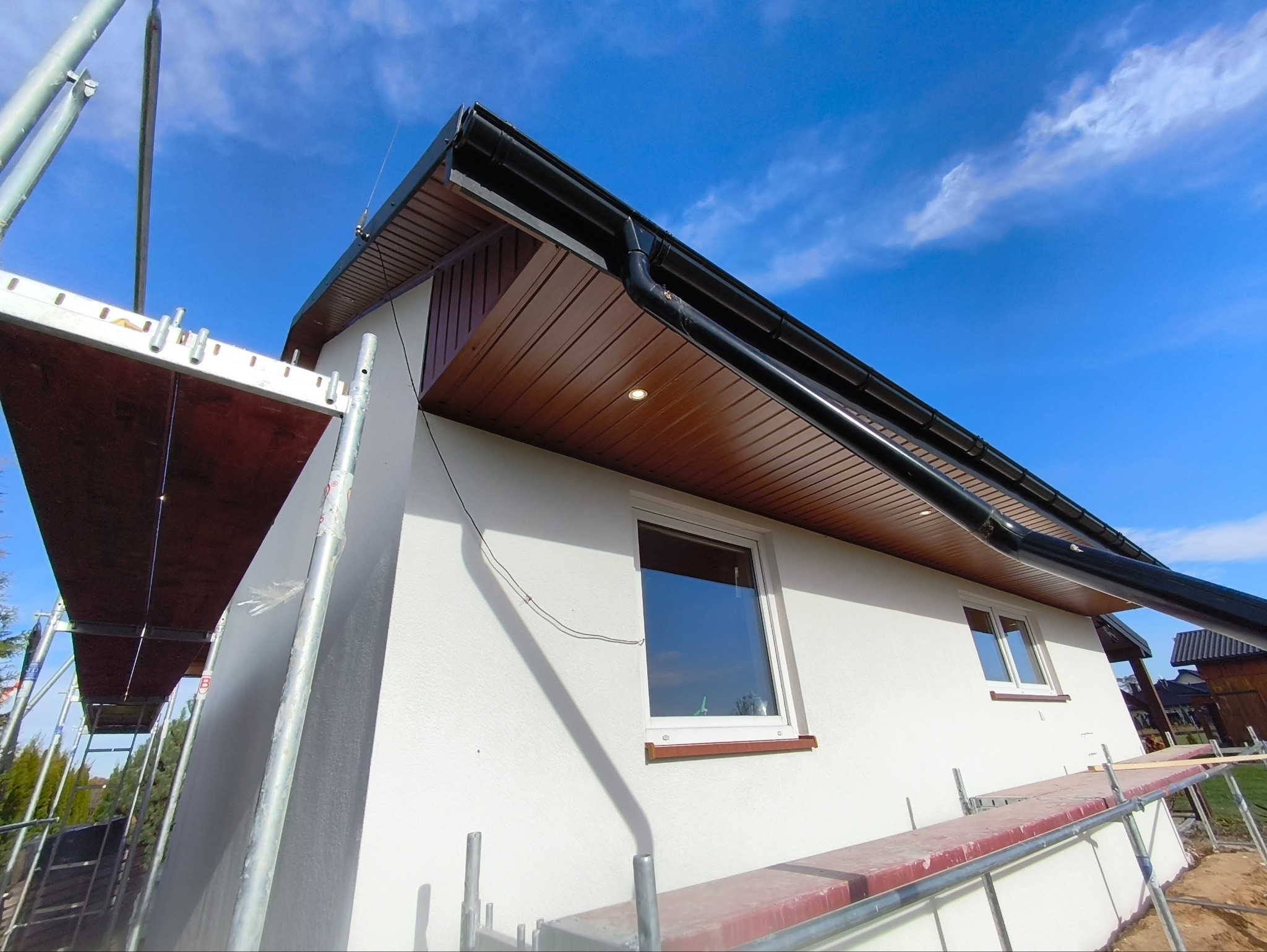 Biała elewacja domu jednorodzinnego z brązową podbitką dachową, widoczna z poziomu rusztowania na tle błękitnego nieba z chmurami. Ujęcie z dołu.