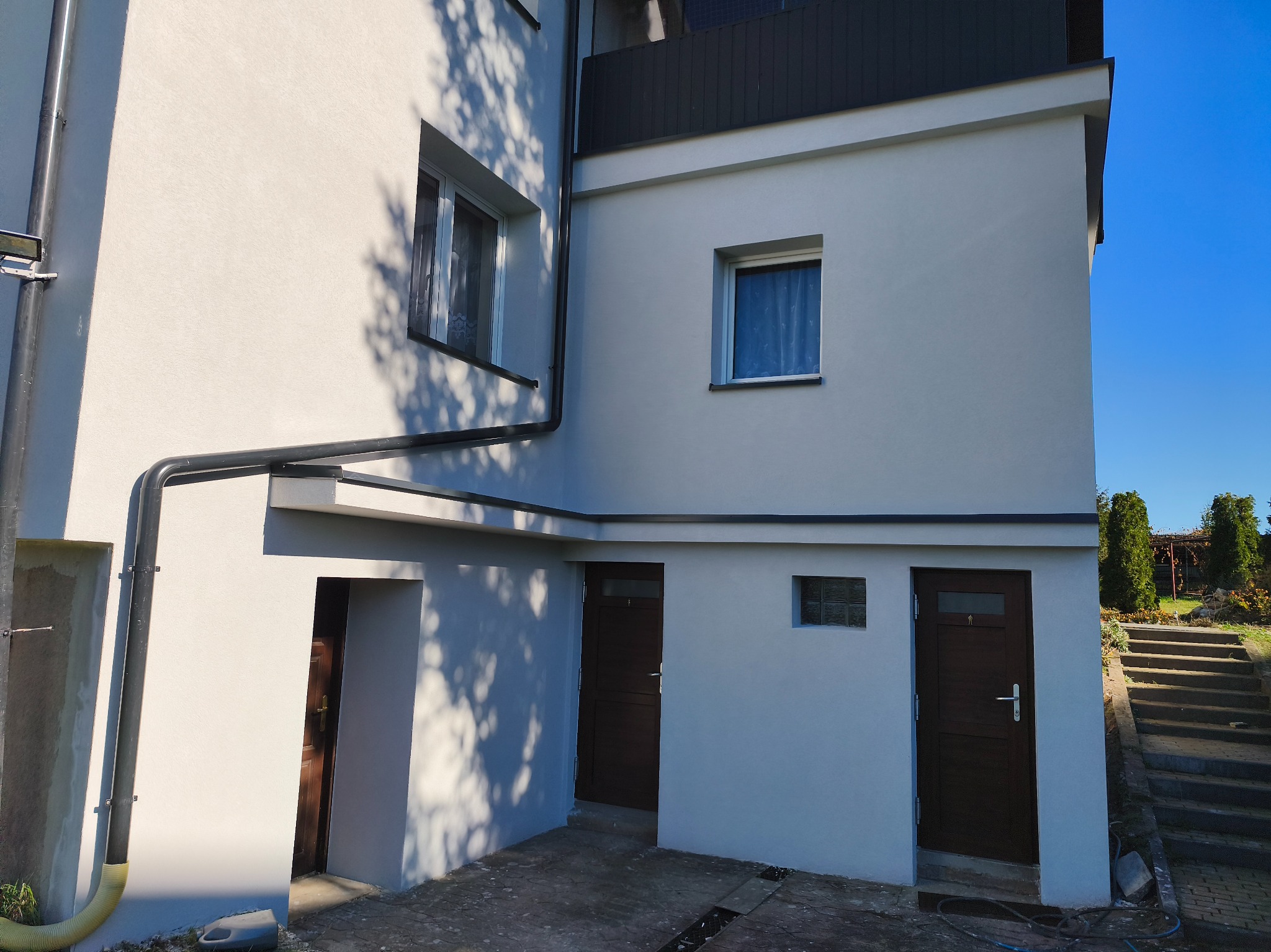 Odnowiona elewacja budynku z nowymi oknami i drzwiami, widoczne detale wykończenia, rynny i schody prowadzące do ogrodu. Jasna kolorystyka i cień drzew na ścianie.