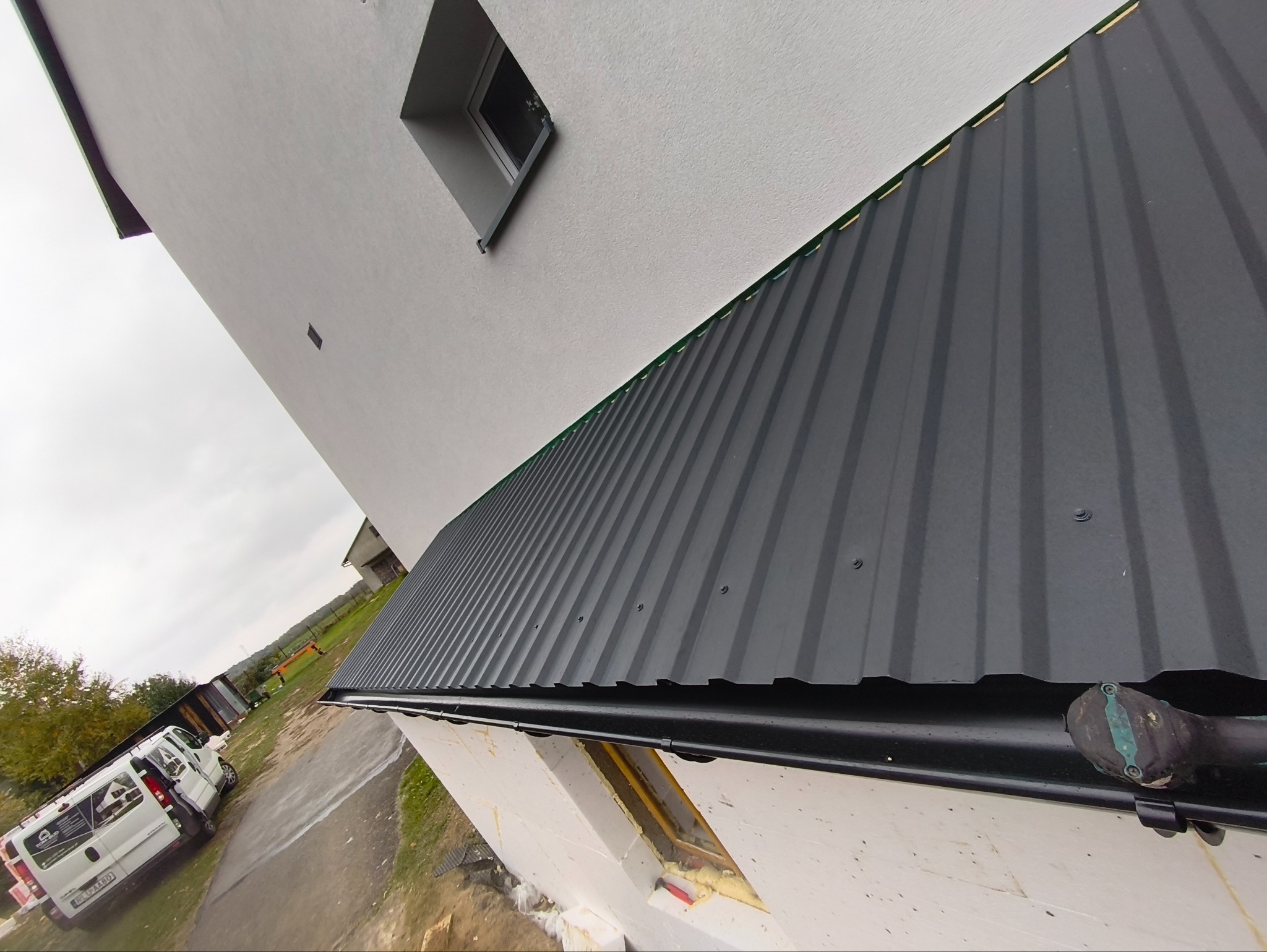 Elewacja budynku z szarym, metalowym dachem i oknem w kształcie rombu. Widoczny fragment ocieplenia ściany i rynna. W tle zaparkowany biały bus.