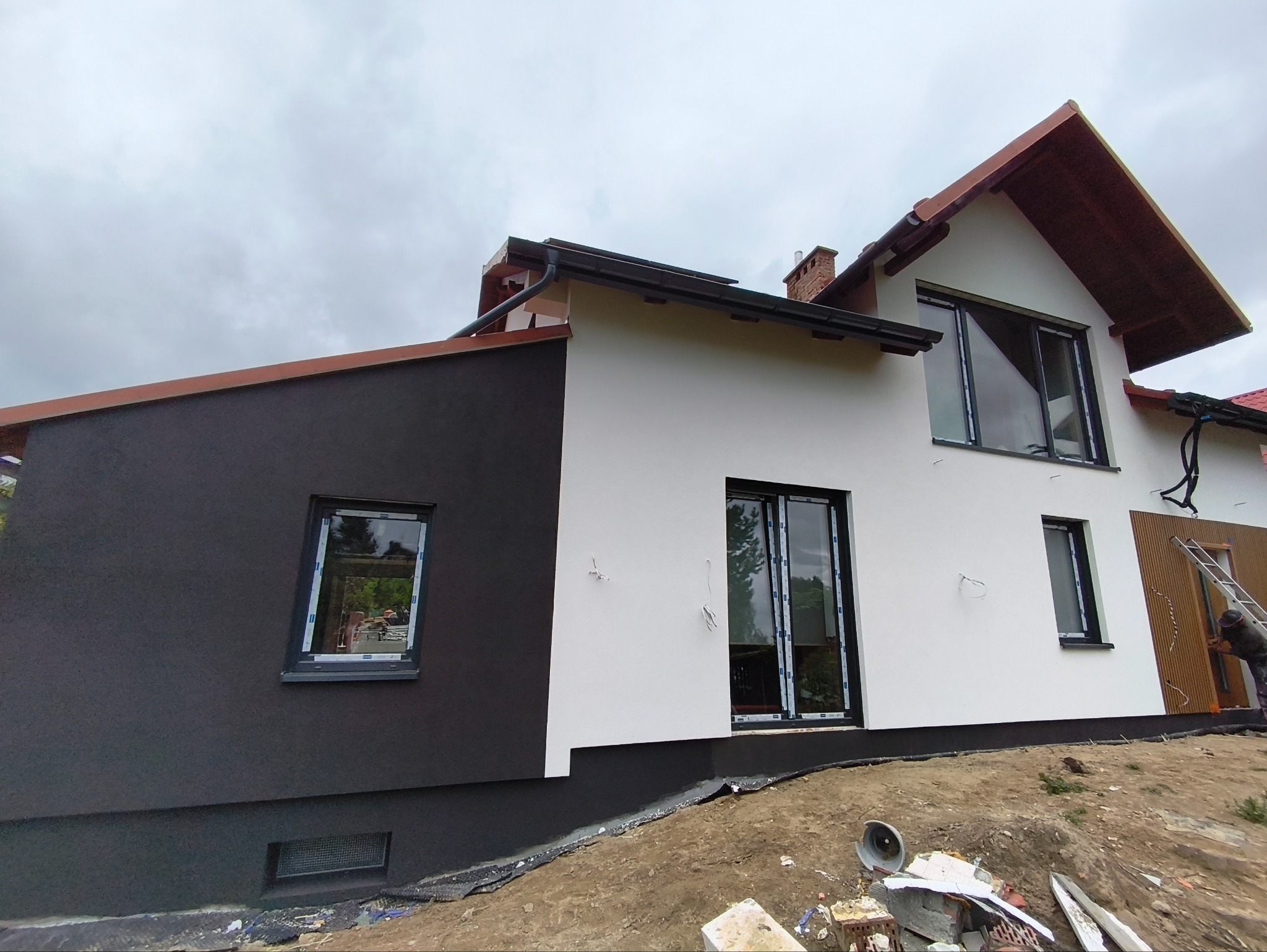Dwukolorowa elewacja domu z czarną i białą częścią. Widoczne okna z czarnymi ramami, drabina i fragment pracownika. Tło: pochmurne niebo.