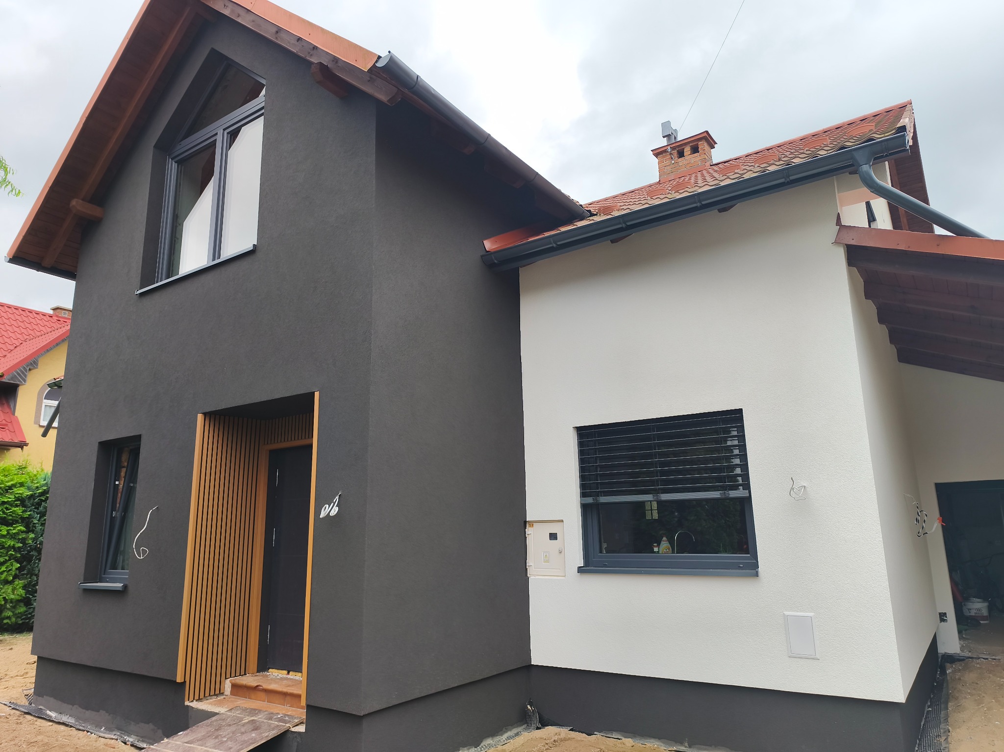 Dwukondygnacyjny dom z elewacją w kolorach grafitu i bieli, z drewnianymi akcentami przy wejściu. Widoczne okna z szarymi ramami i dach z czerwoną dachówką.