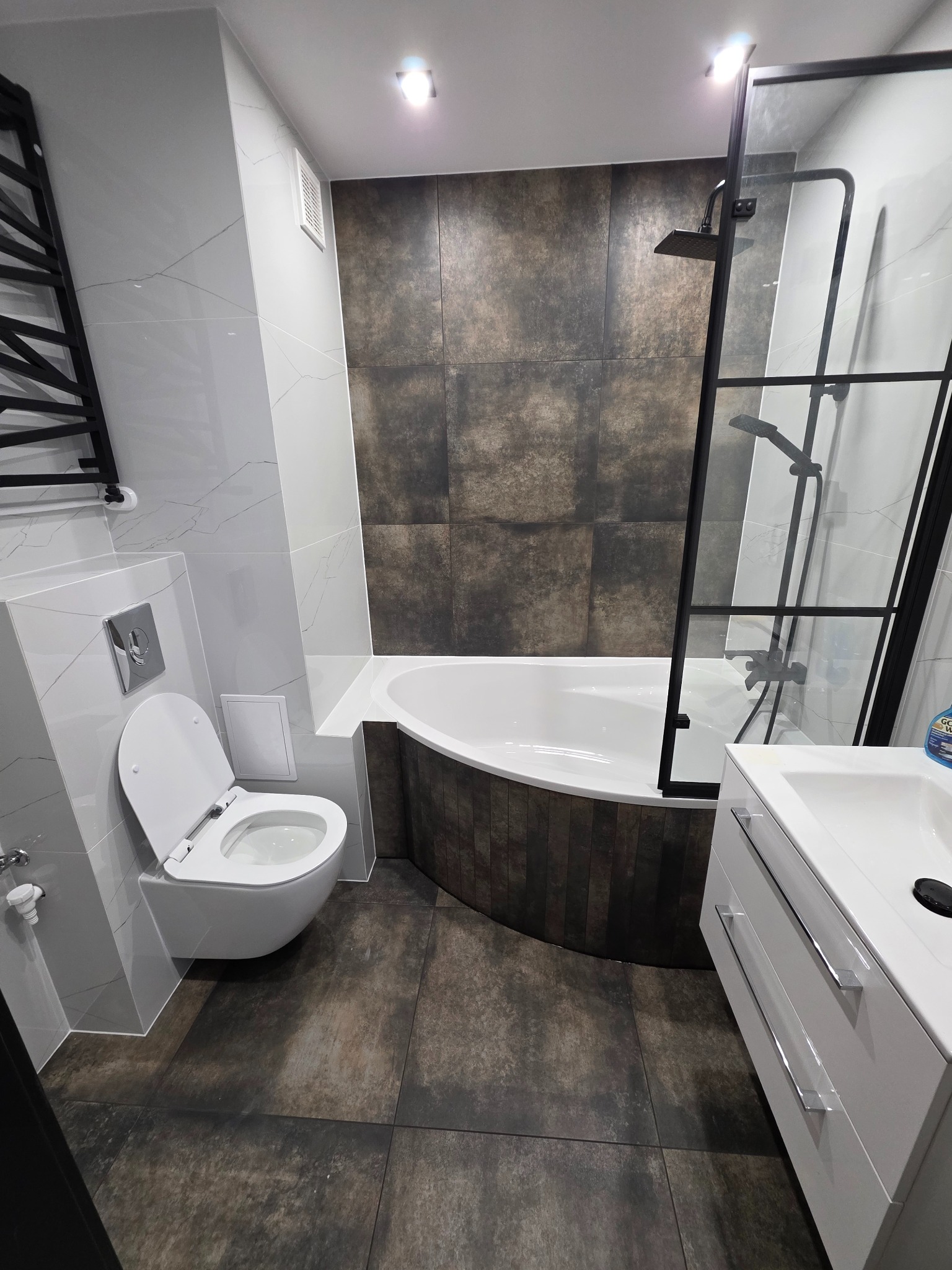 Nowoczesna łazienka po remoncie: biała ceramika, kabina prysznicowa z czarnymi szprosami, brązowe płytki imitujące kamień, minimalistyczna umywalka z szafką.