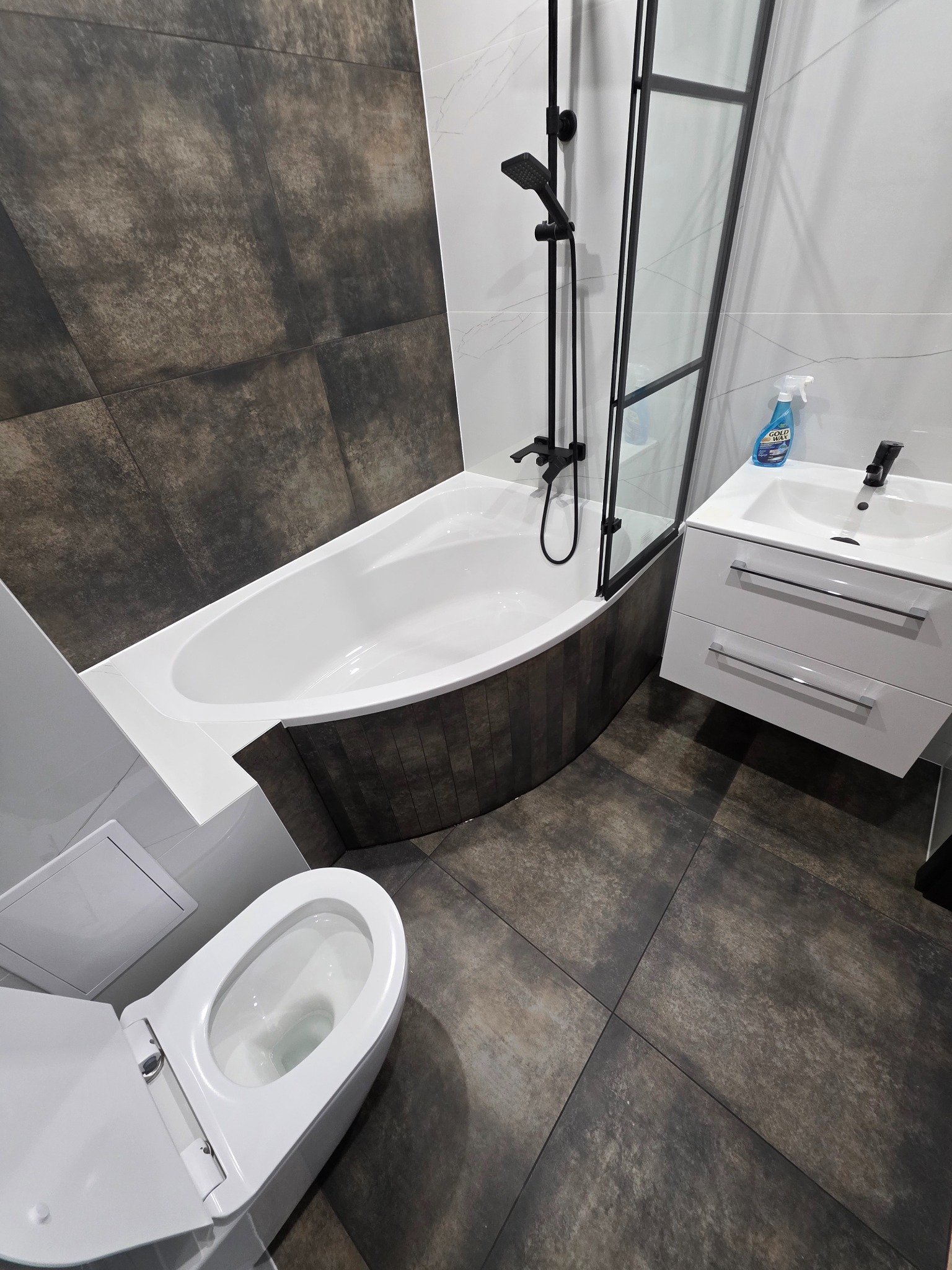 Nowoczesna łazienka z wanną z czarną armaturą, umywalką z szafką i toaletą. Ciemne płytki podłogowe i na ścianie wanny, białe płytki na pozostałych ścianach.