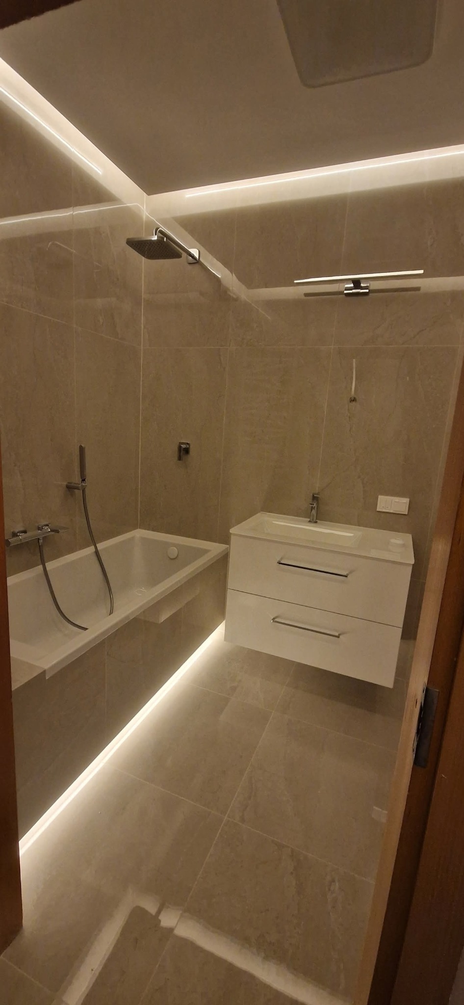 Elegancka łazienka z beżowymi płytkami, podświetleniem LED pod wanną i szafką umywalkową. Nowoczesny design z deszczownicą i minimalistyczną armaturą.