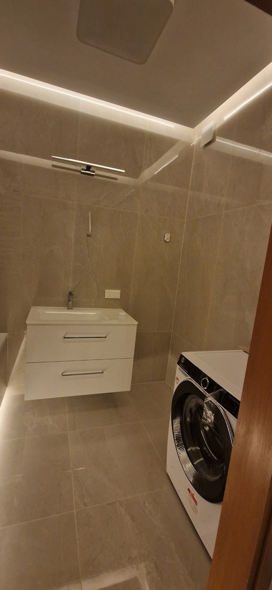 Wykończona łazienka w beżowej tonacji z podświetleniem LED, umywalką z szafką i pralką. Minimalistyczny design, płytki imitujące kamień, nowoczesne oświetlenie.