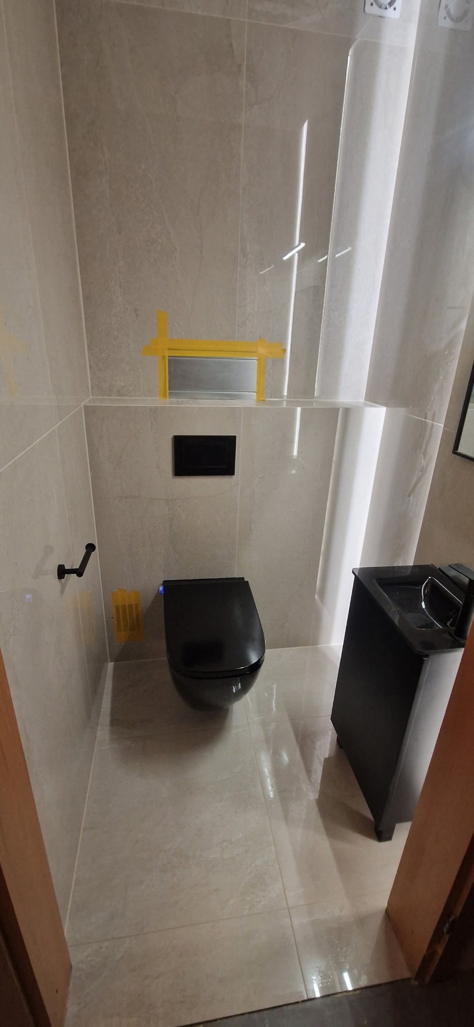 Nowoczesna łazienka z beżowymi płytkami, czarną toaletą podwieszaną i umywalką. Żółta taśma malarska zabezpiecza elementy. Minimalistyczny design.