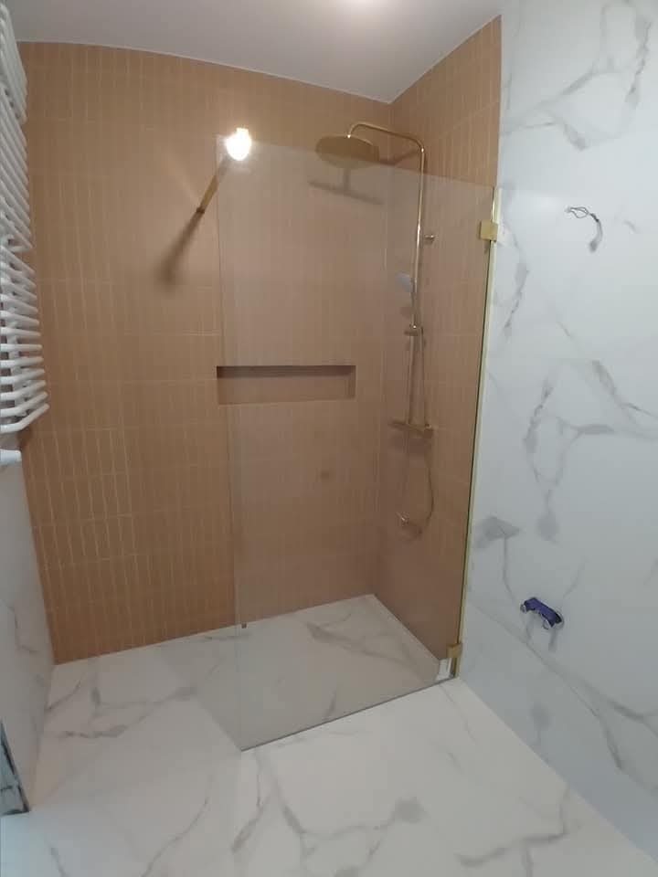 Wykończona łazienka z kabiną prysznicową typu walk-in, beżowe płytki na ścianie i białe marmurowe na podłodze. Złota armatura i szklana ścianka kabiny.