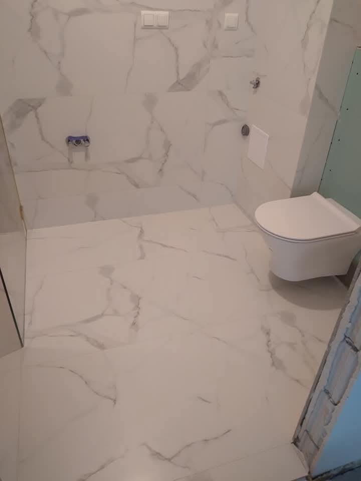 Biała łazienka z marmurowymi płytkami, wisząca toaleta i fragment ściany z płyt karton-gips, widoczne elementy instalacji wodnej. Minimalistyczny design.