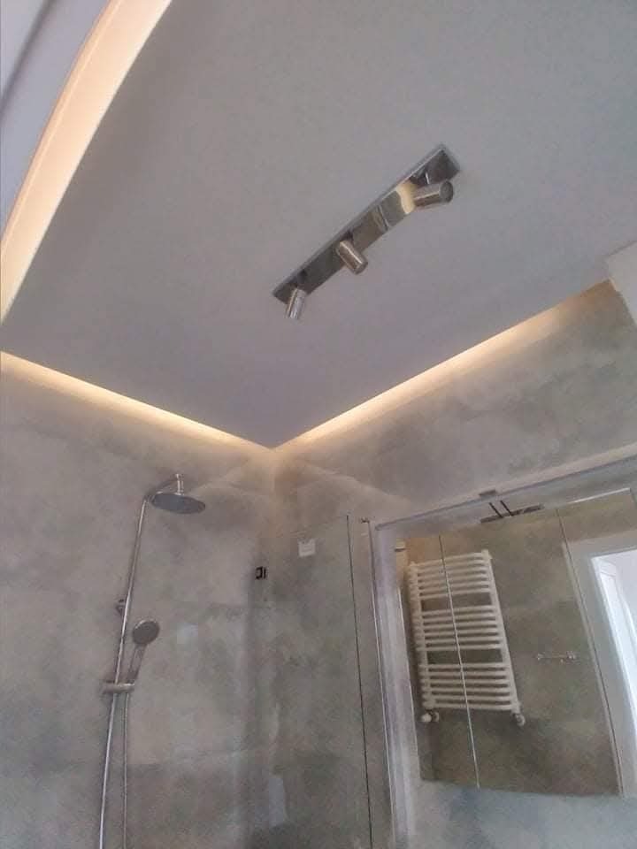 Nowoczesna łazienka w Warszawie: szara ściana z efektem betonu, oświetlenie LED podsufitowe i punktowe, kabina prysznicowa z grzejnikiem drabinkowym.