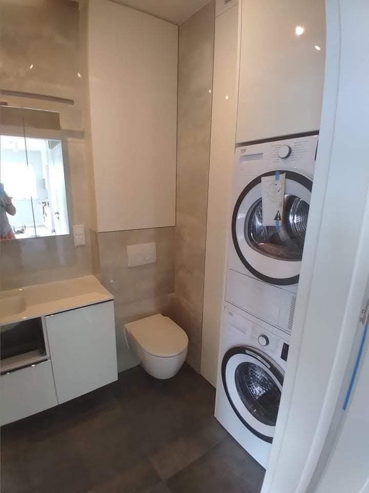 Nowoczesna łazienka z pralką i suszarką w zabudowie, umywalką z szafką i podwieszaną toaletą. Minimalistyczny design, płytki imitujące kamień, oświetlenie punktowe.