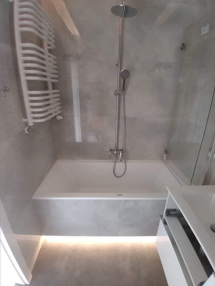 Nowoczesna łazienka z wanną, deszczownicą i grzejnikiem drabinkowym. Szare płytki, subtelne oświetlenie LED pod wanną. Minimalistyczny design.