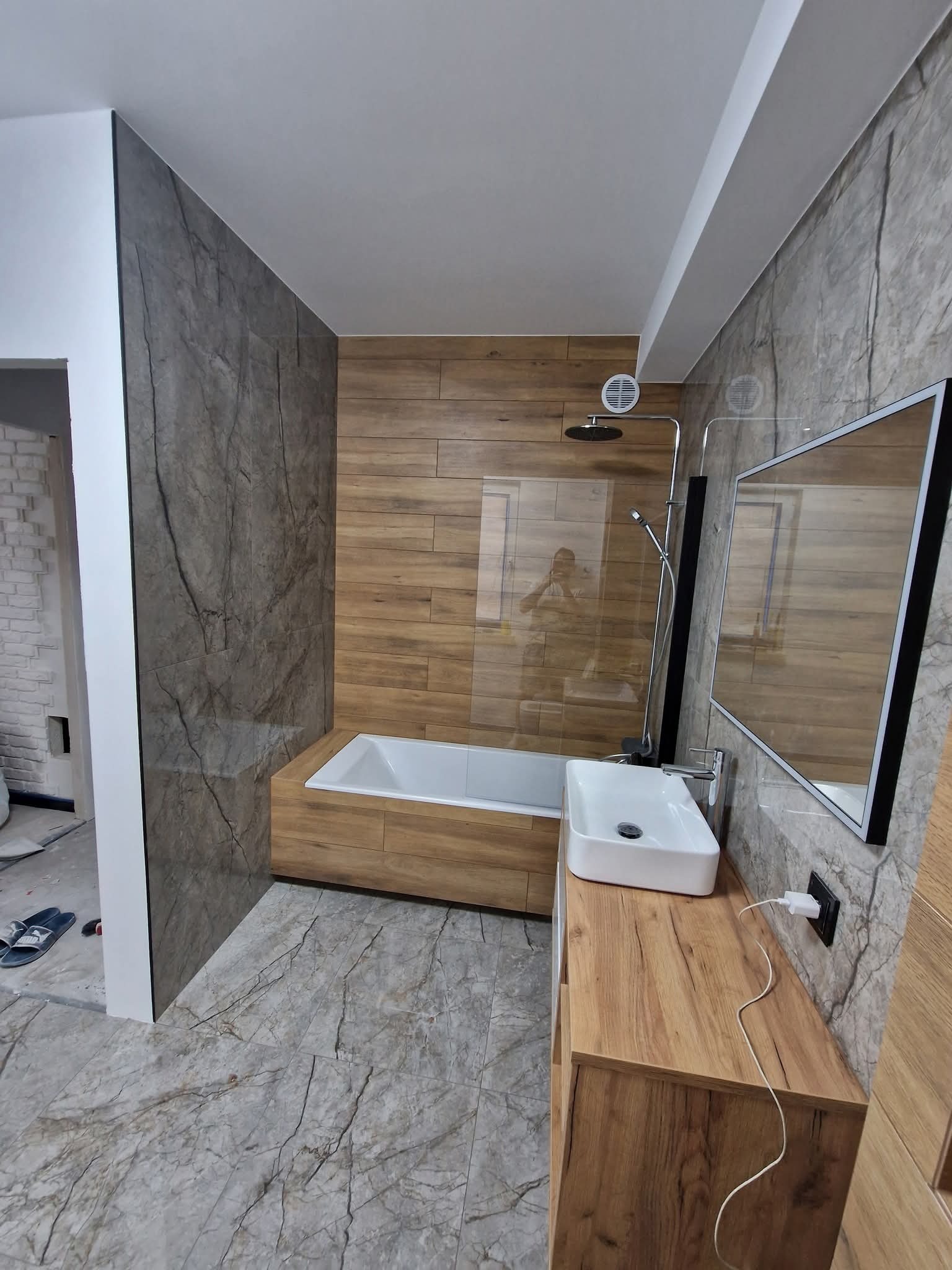 Nowoczesna łazienka po remoncie: drewnopodobne płytki, marmurowe ściany, biała umywalka i wanna. Widoczne odbicie osoby w szklanej ściance prysznica.