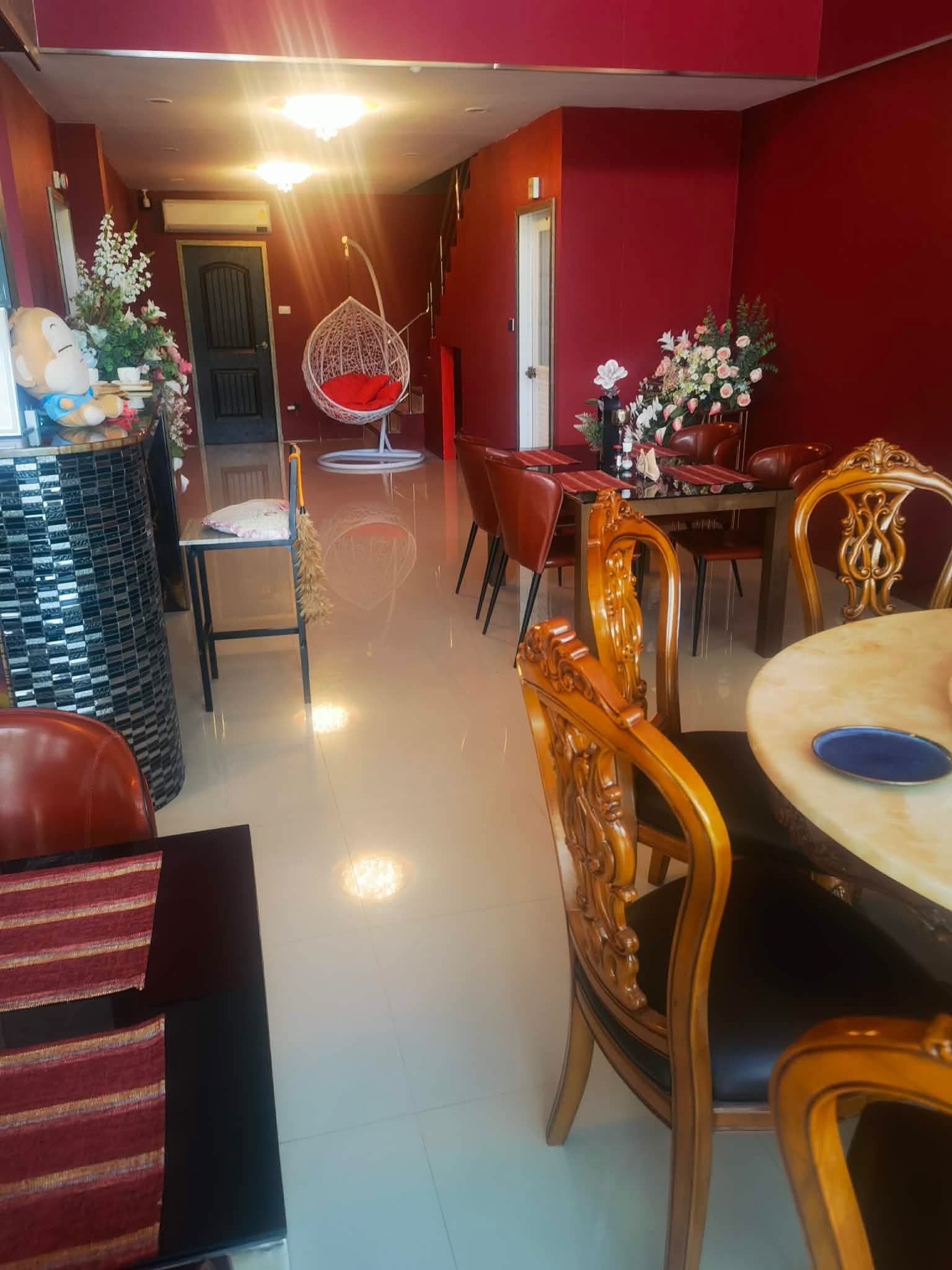 Eleganckie wnętrze jadalni z czerwonymi ścianami, ozdobnymi krzesłami i wiszącym fotelem. Widoczne kwiaty i dekoracje. Jasne oświetlenie.