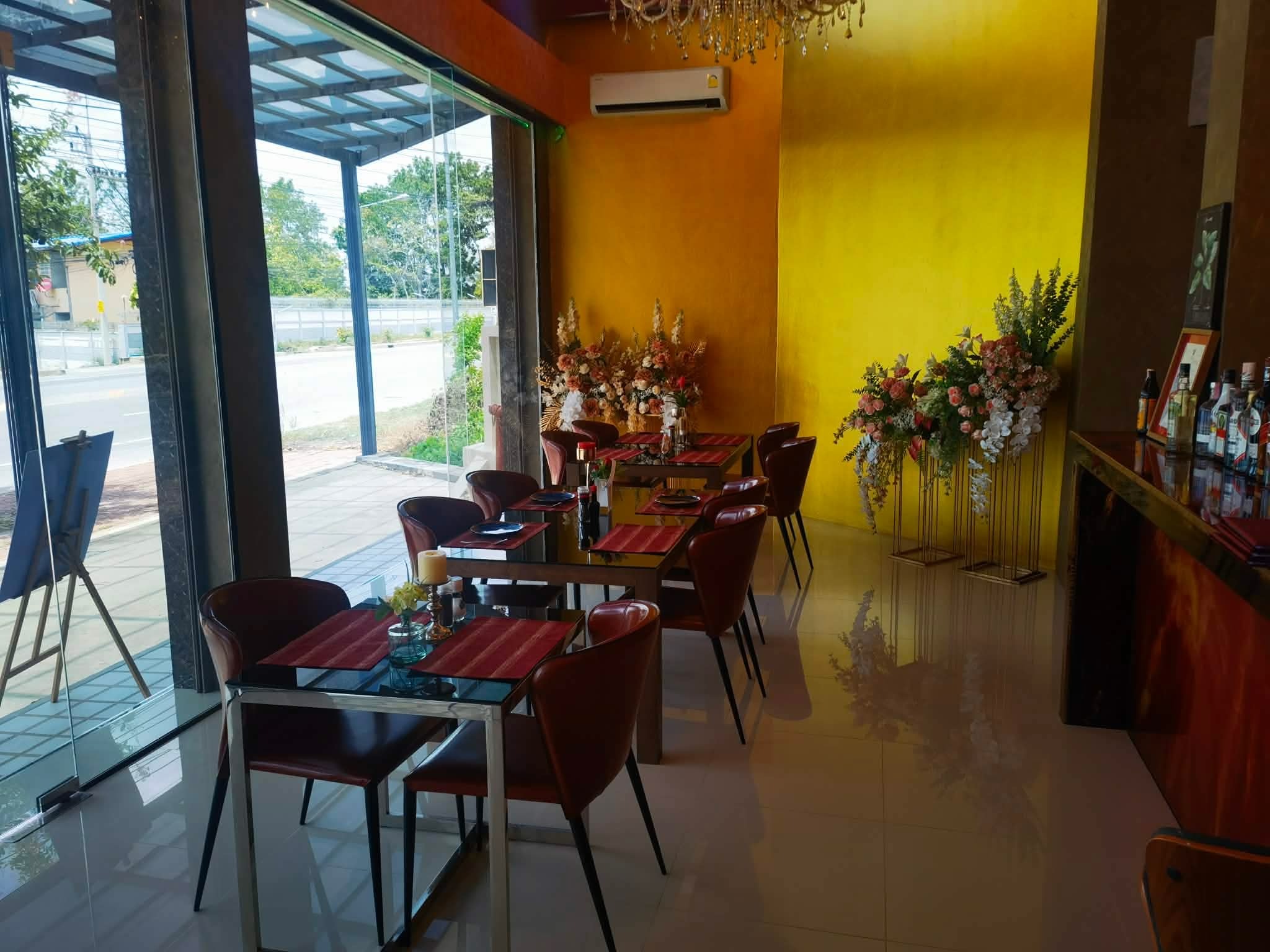 Eleganckie wnętrze restauracji z żółtą ścianą, stolikami nakrytymi do posiłku i dekoracjami kwiatowymi. Widoczne krzesła z brązowej skóry i szklane elementy.