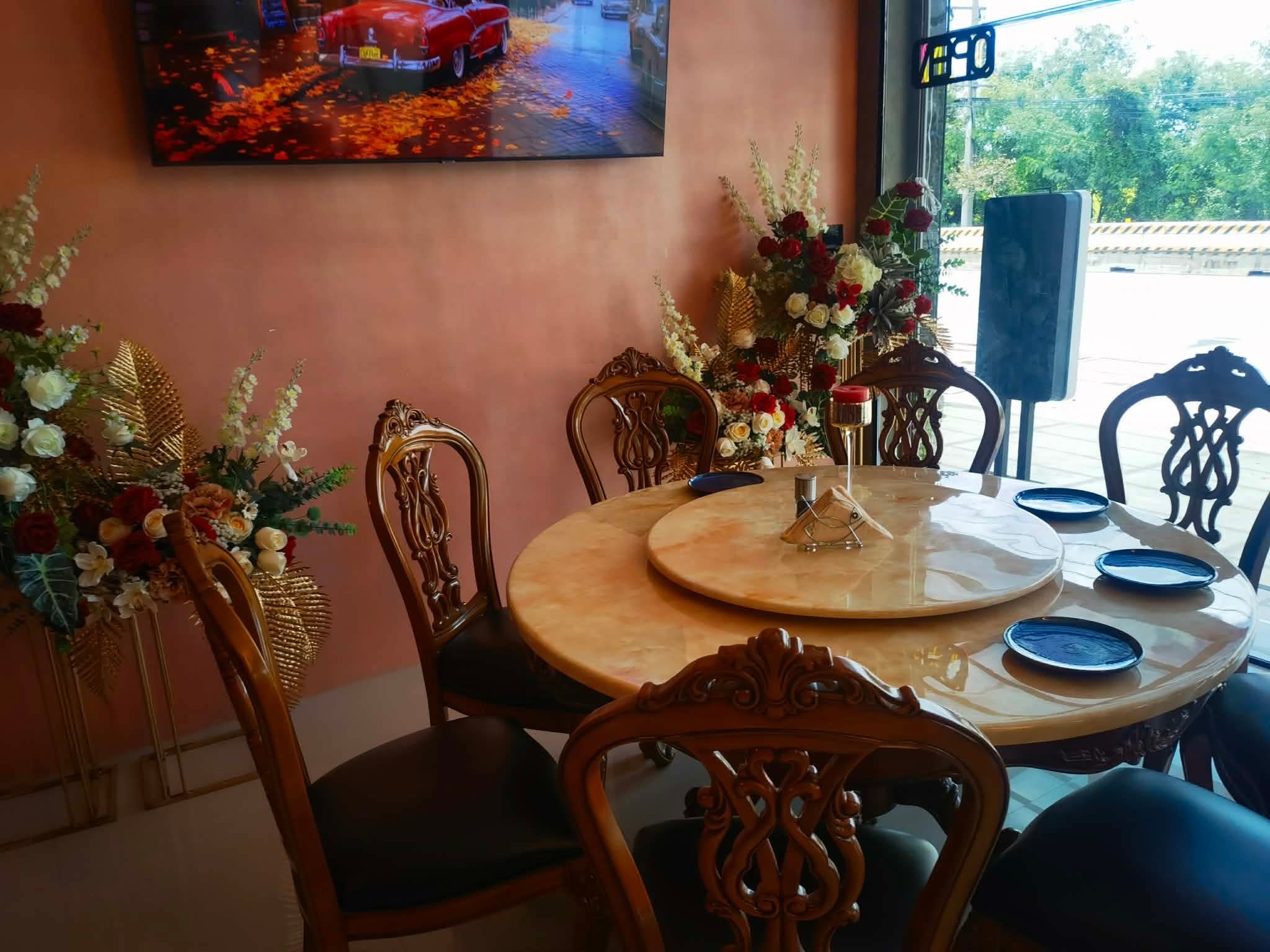 Elegancka jadalnia z okrągłym stołem z marmurowym blatem, otoczonym rzeźbionymi krzesłami. Na ścianie telewizor z obrazem czerwonego samochodu. Dekoracje kwiatowe dodają uroku.