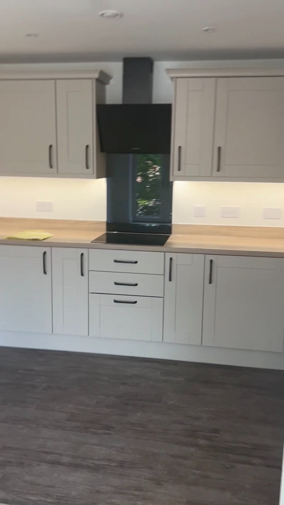 Jasna, nowoczesna kuchnia z beżowymi szafkami, drewnianym blatem i czarnym okapem. Widoczne oświetlenie podszafkowe i ciemna podłoga. Minimalistyczny design.