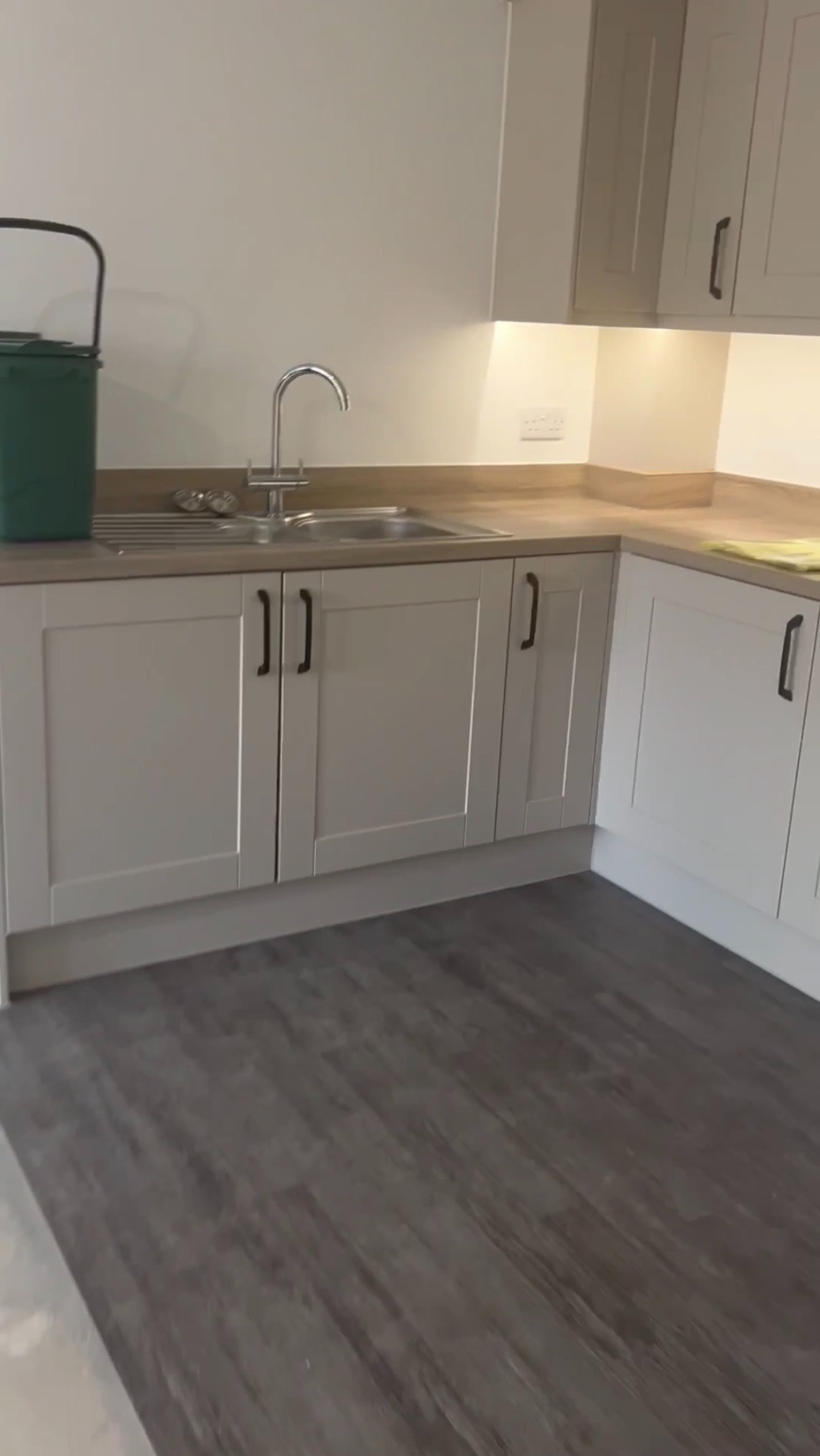 Jasna kuchnia z szarymi szafkami i drewnianym blatem. Ujęcie na zlew i fragment podłogi imitującej drewno. Minimalistyczny design, nowoczesne wykończenie.