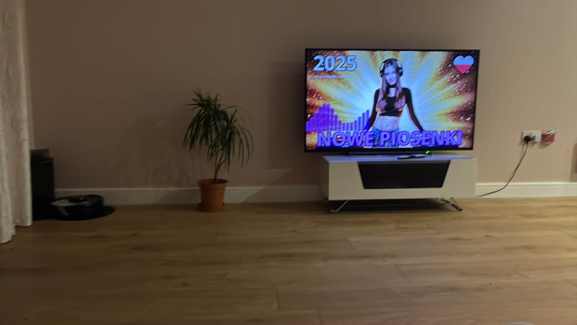 Salon z parkietem: telewizor z napisem 'Nowe Piosenki', roślina w doniczce, robot sprzątający. Minimalistyczny wystrój, ciepłe oświetlenie.