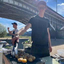 FoodPassionMan Jarosław Okrzejski - Kucharz w czarnym fartuchu grilluje ziemniaki pod mostem. Drugi kucharz w tle. Plaża, błękitne niebo. Nietypowa lokalizacja cateringu.