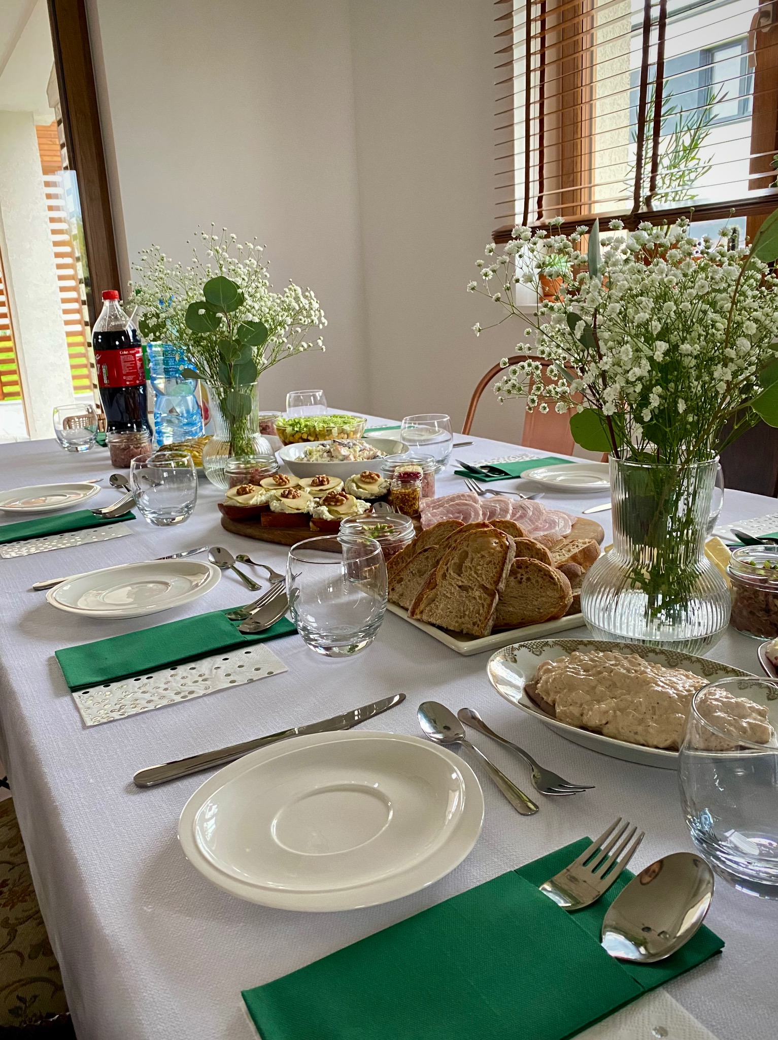 Uroczysty stół z białym obrusem, zielonymi serwetkami, kwiatami gipsówki w wazonach i różnorodnymi potrawami, gotowy na przyjęcie gości w jasnym wnętrzu.
