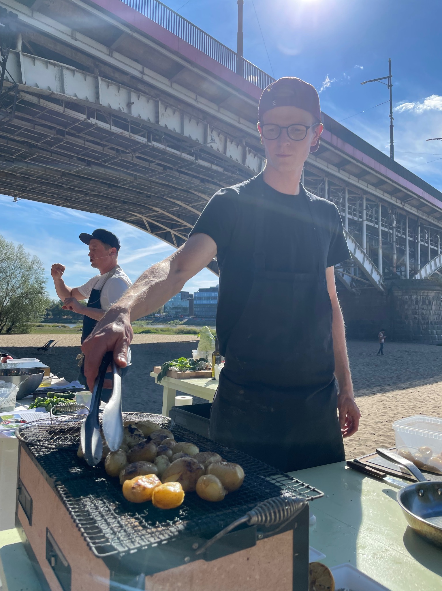 Kucharz w czarnym fartuchu grilluje ziemniaki pod mostem. Drugi kucharz w tle. Plaża, błękitne niebo. Nietypowa lokalizacja cateringu.