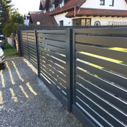 SPAWPAW - Nowoczesne, poziome przęsła ogrodzeniowe w kolorze grafitowym, z widoczną strukturą drewna, na tle domu z czerwoną dachówką i żwirowym podłożem. Słoneczny dzień.