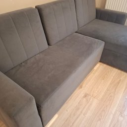 Fabryka Świeżości - Szara, narożna kanapa z welurową tapicerką na drewnianej podłodze. Widoczne pionowe przeszycia na oparciach i delikatne ślady użytkowania na siedzisku.