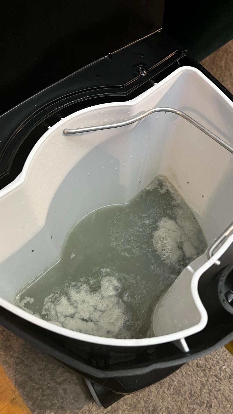 Brudna woda po czyszczeniu w zbiorniku maszyny do prania tapicerki, widoczne drobinki i piana. Zbiornik w kolorze białym, maszyna czarna.