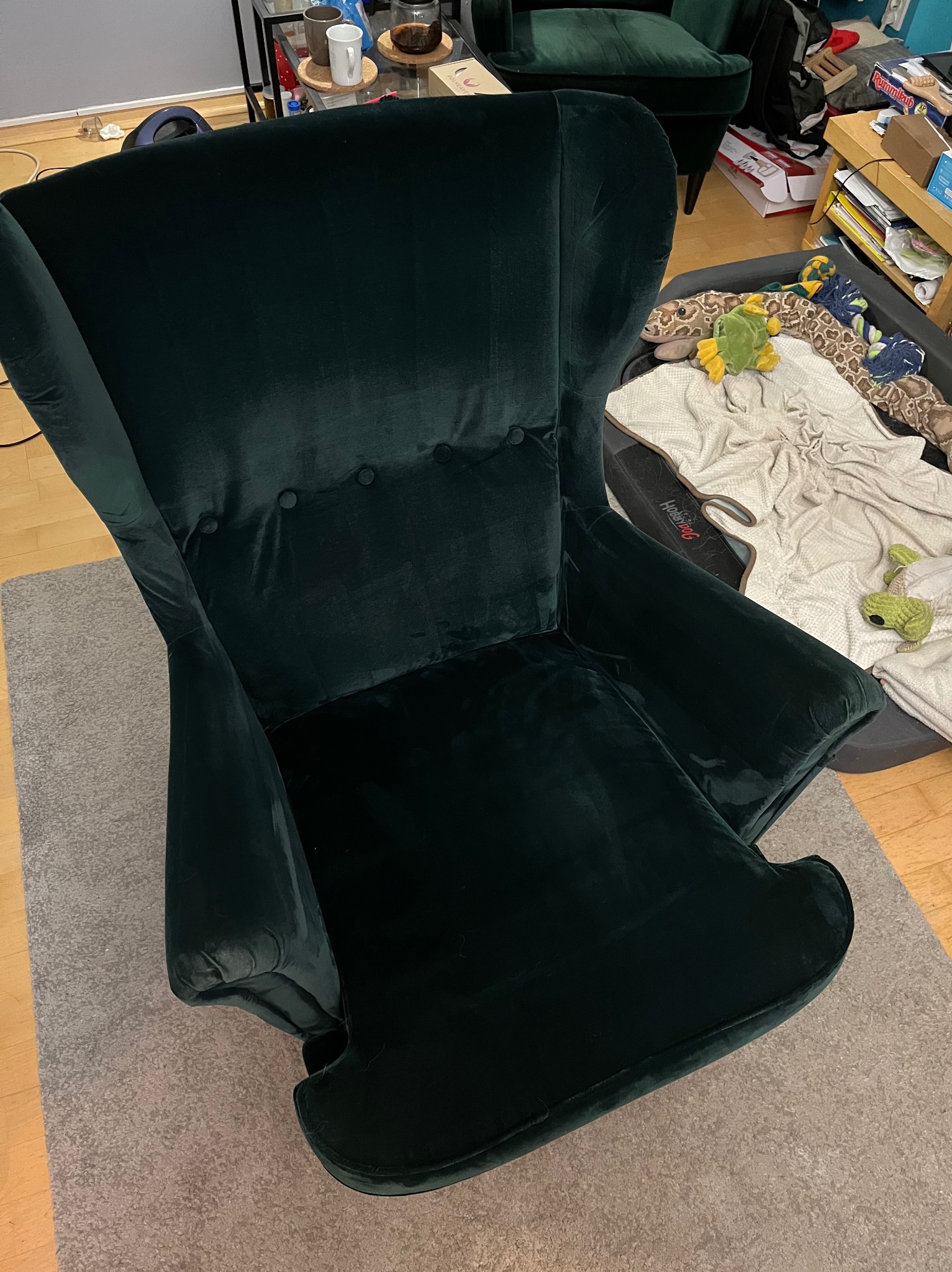 Głęboki, szmaragdowy fotel uszak z aksamitną tapicerką, widoczny z góry na tle szarego dywanu i dziecięcego kojca. Widoczne detale: guziki na oparciu, charakterystyczny kształt.