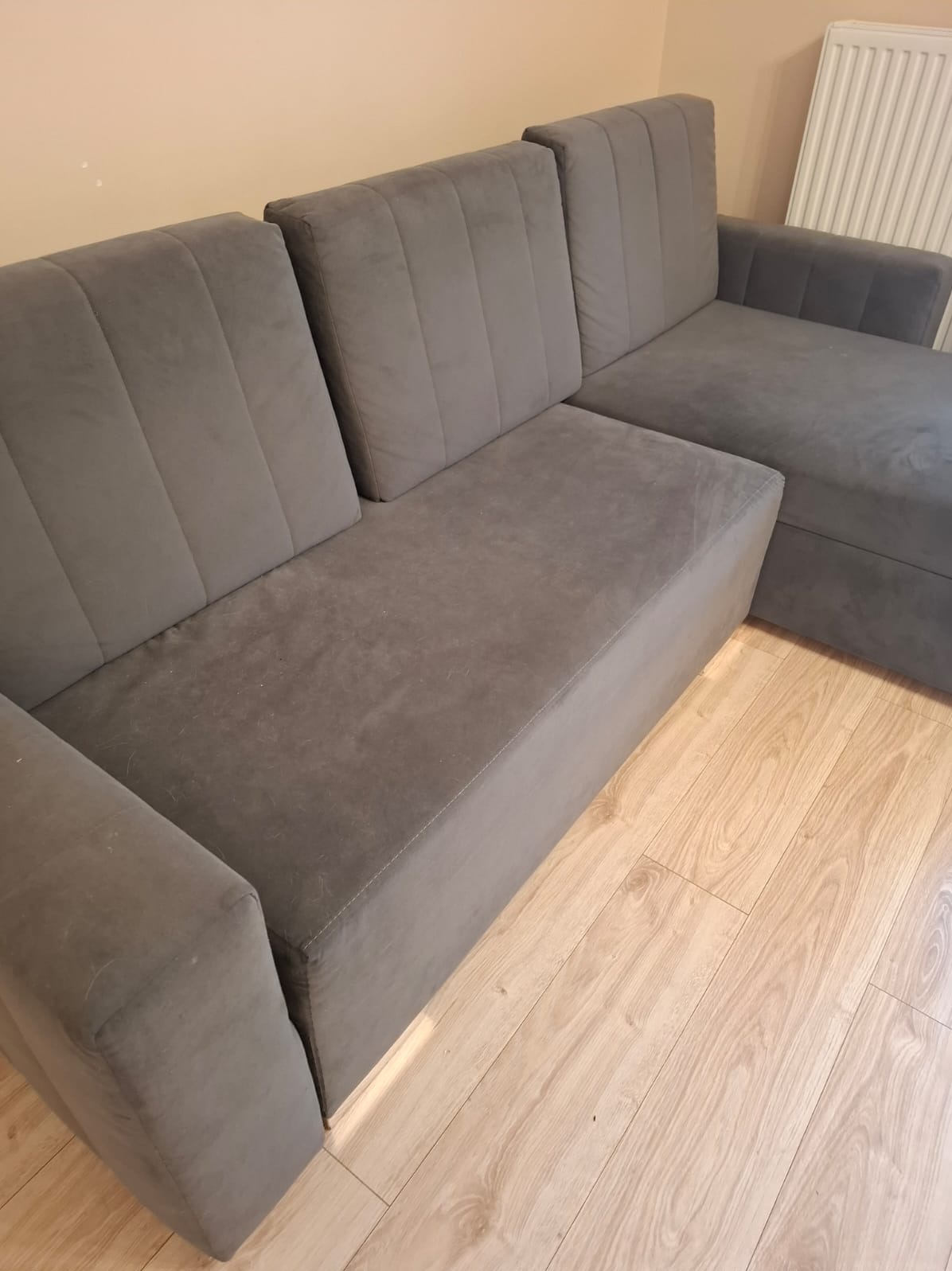 Szara, narożna kanapa z welurową tapicerką na drewnianej podłodze. Widoczne pionowe przeszycia na oparciach i delikatne ślady użytkowania na siedzisku.