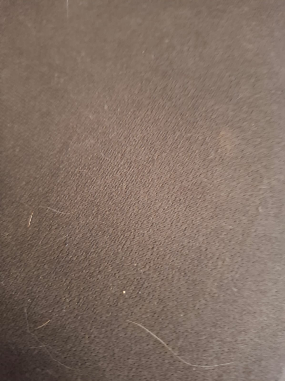 Zbliżenie na brązową, teksturowaną tkaninę tapicerską z widocznymi drobnymi zabrudzeniami i pojedynczymi włosami. Delikatne światło podkreśla strukturę materiału.