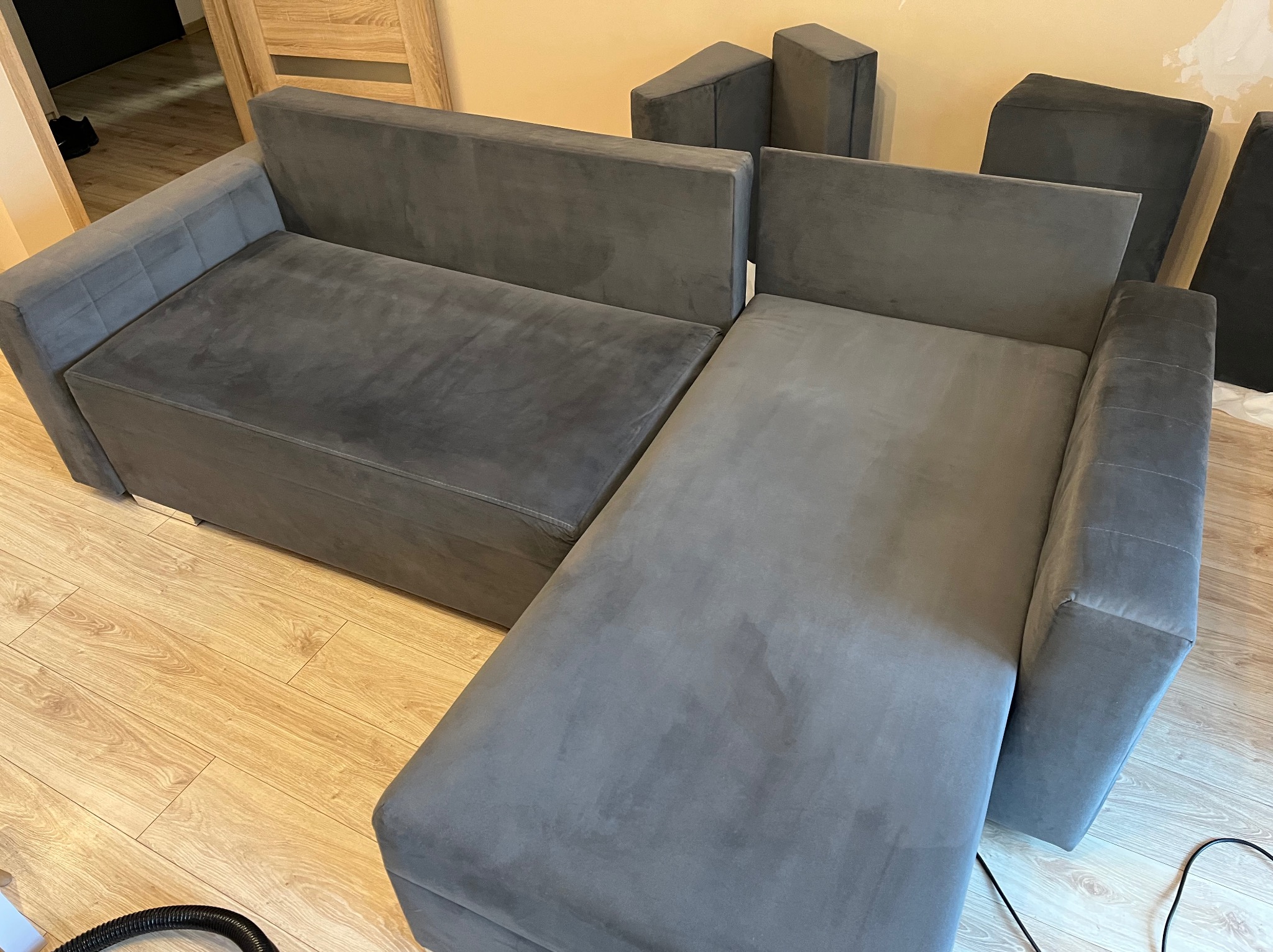 Szara, rozłożona sofa modułowa z widocznymi plamami na tapicerce, sugerująca potrzebę czyszczenia. Na podłodze widoczny wąż odkurzacza.