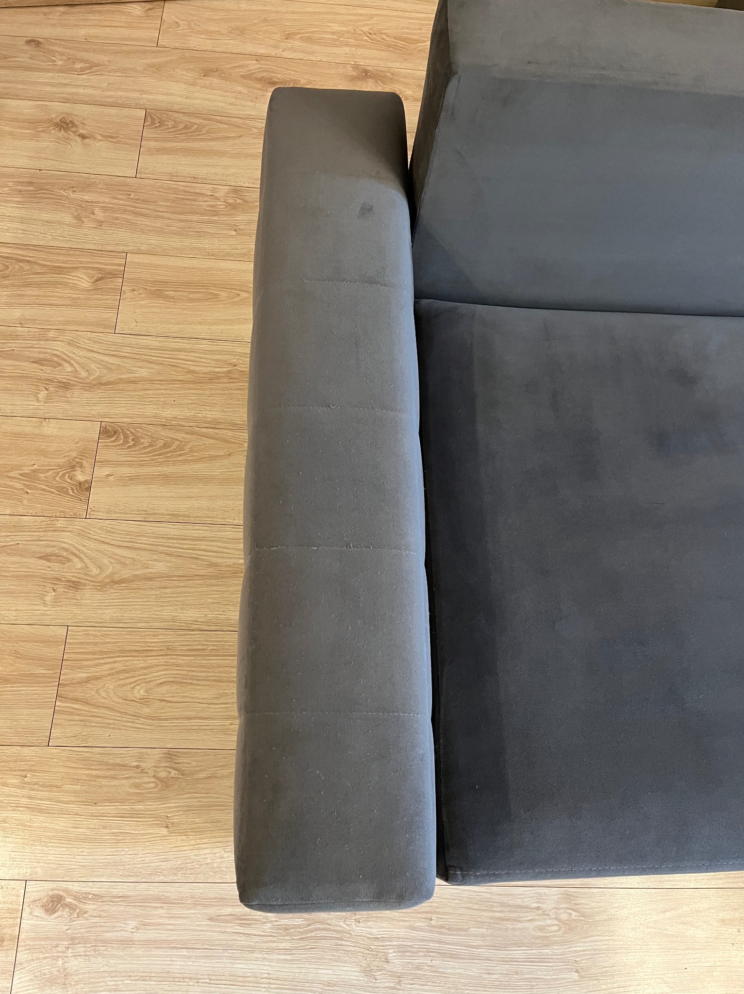 Fragment szarej, welurowej kanapy na drewnianej podłodze. Widoczny bok i siedzisko, ukazujące fakturę materiału i detale wykończenia.