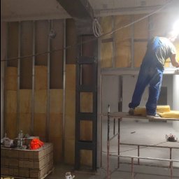 Marulbut - Prace wykończeniowe: montaż stelaża pod płyty gipsowo-kartonowe. Widoczny pracownik na rusztowaniu, metalowy stelaż, izolacja z wełny mineralnej i materiały budowlane.