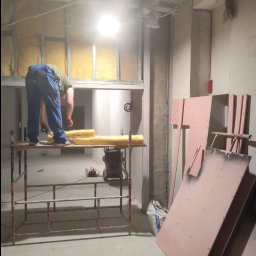 Marulbut - Pracownik na rusztowaniu montuje izolację z wełny mineralnej w metalowym stelażu ściany. W tle widoczne płyty kartonowo-gipsowe i surowy stan wykończenia wnętrza.