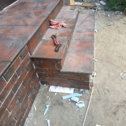 Marulbut - Wykończone płytkami schody zewnętrzne z ceglanym obrzeżem. Na drugim stopniu leżą narzędzia: szczotka i nóż. W tle piasek i resztki materiałów budowlanych.