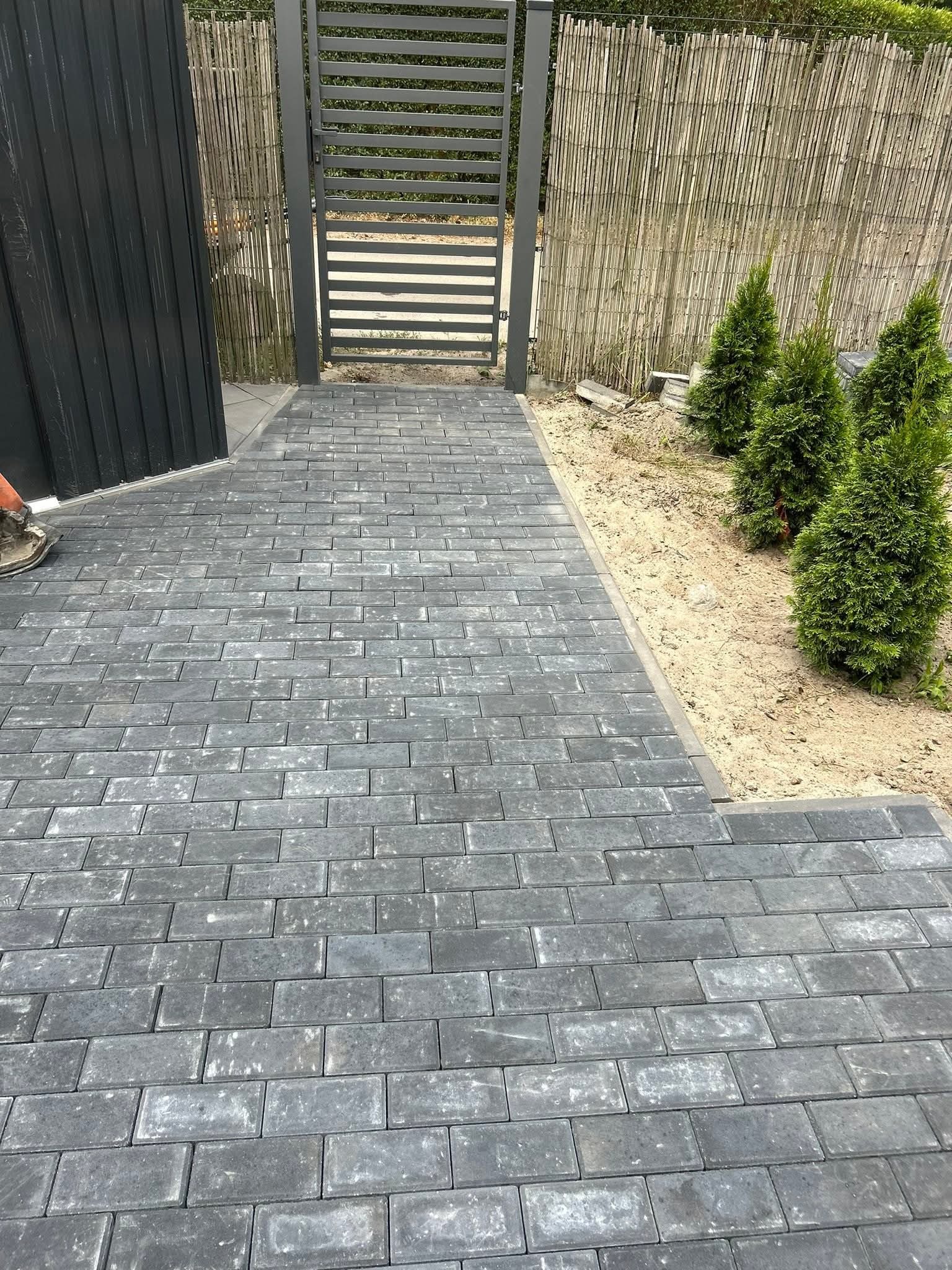 Ułożona szara kostka brukowa prowadząca do metalowej furtki, obok rabata z tujami i bambusowy płot. Widoczny fragment buta pracownika.
