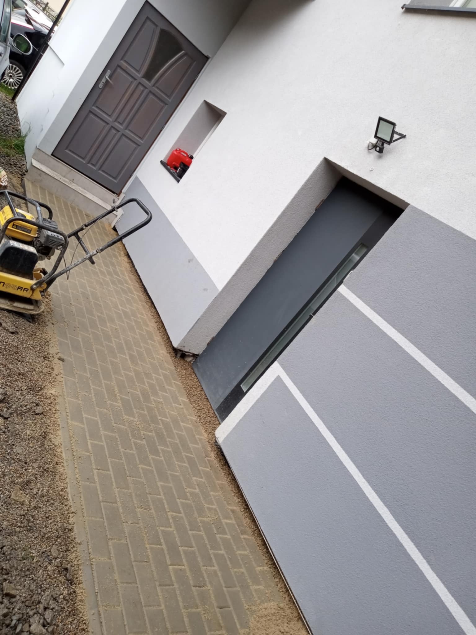 Ułożona kostka brukowa w jodełkę przy budynku z szarą elewacją i ubijarką. Widoczny fragment samochodu i drzwi wejściowe.