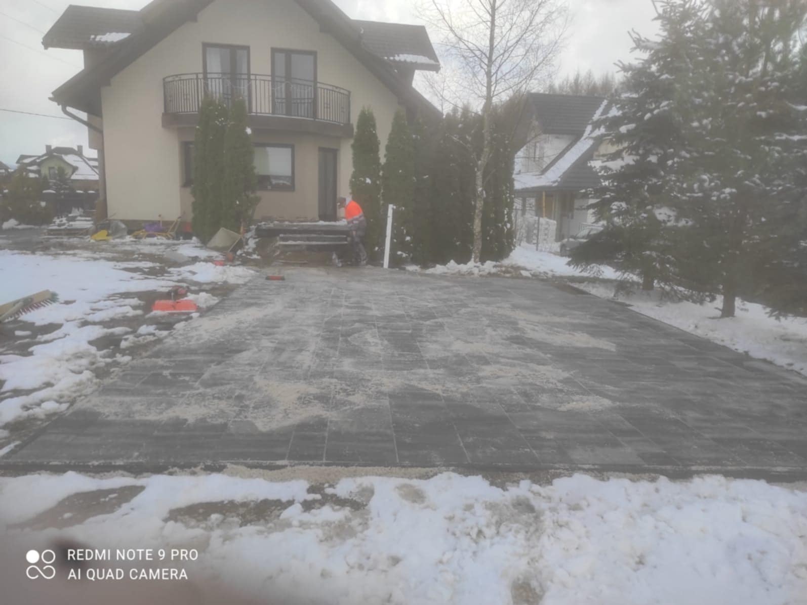 Ułożona kostka brukowa na posesji w zimowej scenerii. Widoczny fragment domu z balkonem i pracownik w odblaskowej kamizelce. Częściowo zasypane śniegiem.