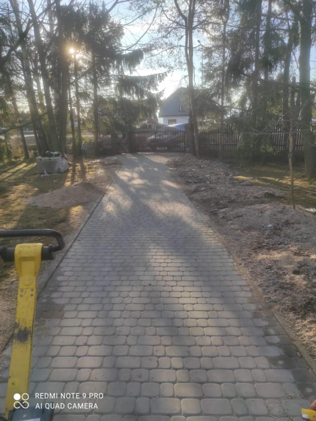Ułożona ścieżka z kostki brukowej prowadząca do bramy, z żółtą zagęszczarką w lewym dolnym rogu. W tle drzewa i dom.