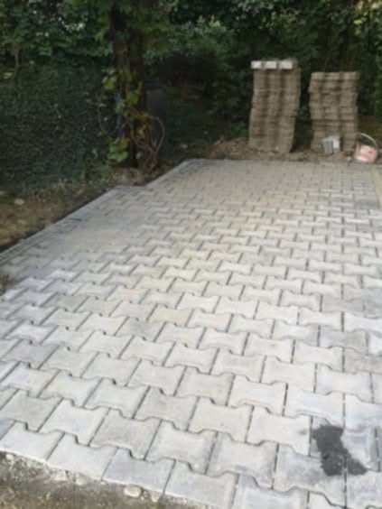 Ułożona, szara kostka brukowa typu 'I' na podwórku, z dwoma stosami kostki w tle. Widoczne drzewa i zieleń. Częściowo widoczny kubeł budowlany.