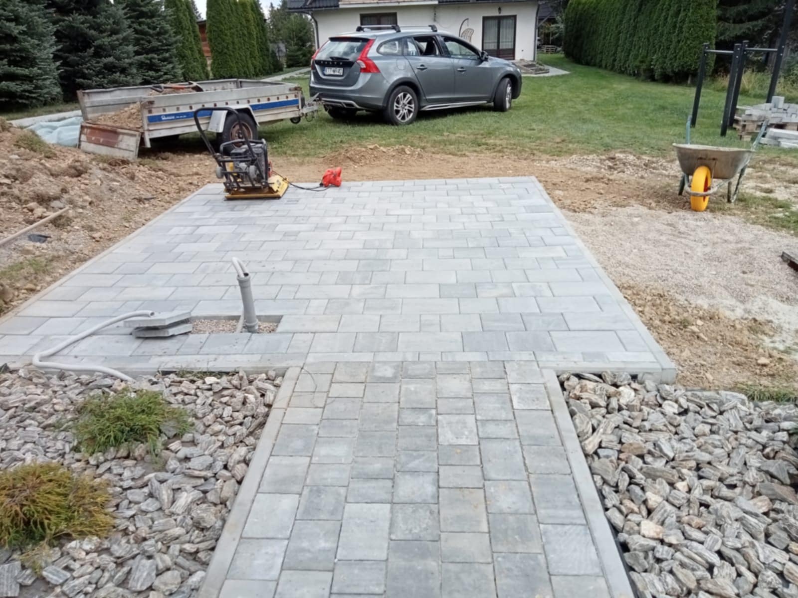 Ułożona szara kostka brukowa na posesji z widocznym przyłączem wodnym, ubijarką, przyczepą i samochodem w tle. Wykończone obrzeża z kamieni ozdobnych.