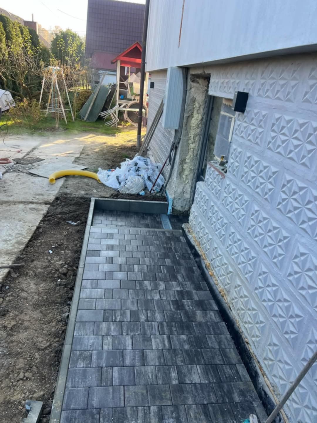 Ułożona, szara kostka brukowa przy ścianie budynku z dekoracyjną fakturą. Widoczne przygotowane podłoże i elementy otoczenia, w tym rura drenażowa.