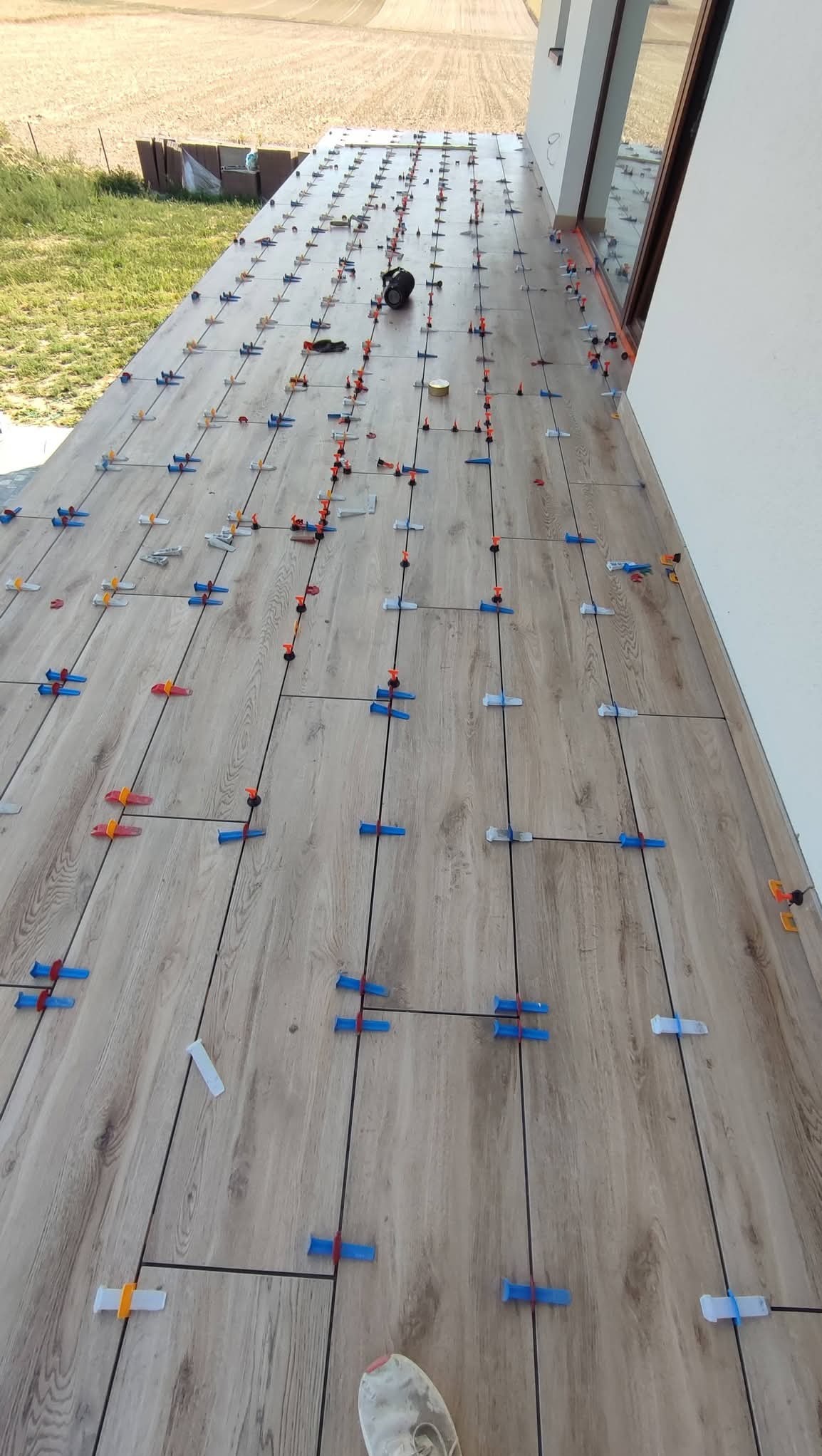 Kafelki imitujące drewno na tarasie, w trakcie montażu z użyciem systemu poziomowania. Widoczne krzyżyki dystansowe i kliny. W tle trawa i budynek.