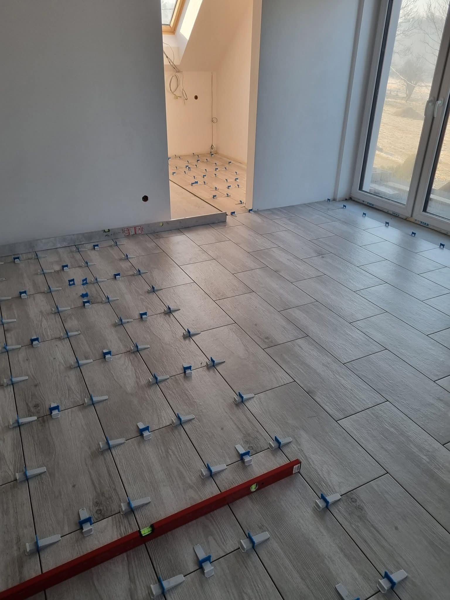 Ułożone płytki podłogowe imitujące drewno, z krzyżykami dystansowymi i poziomicą w pomieszczeniu z oknem i widokiem na zewnątrz. Częściowo ułożona podłoga w jasnym wnętrzu.