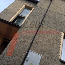 FULGUR GRZEGORZ WILKOSZ - Zewnętrzna instalacja elektryczna na elewacji budynku z szarą skrzynką i czarnym kablem w peszlu, mocowanym klamrami. Widok z dołu, lekki kąt.